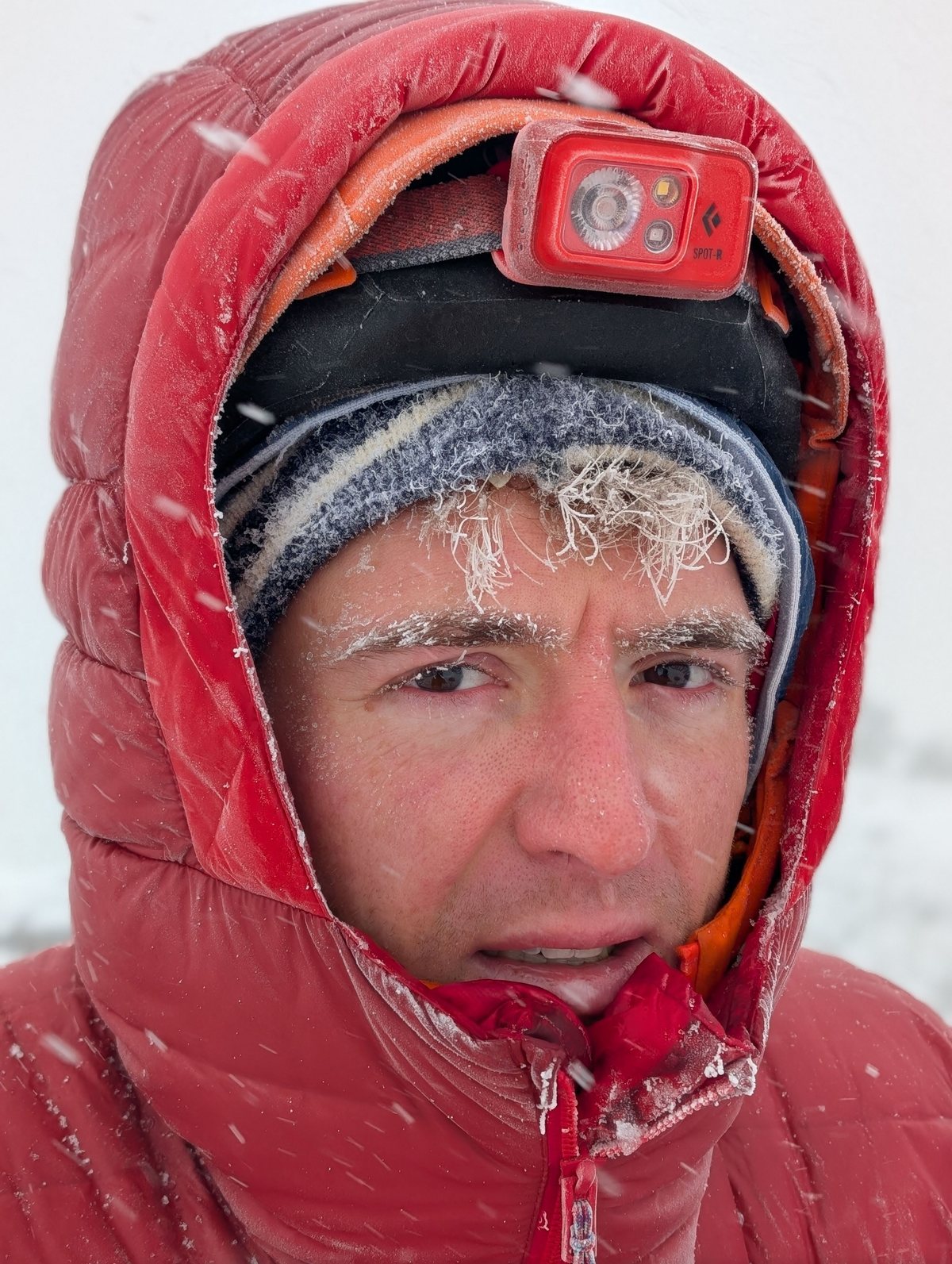 Sam in a red down hood at altitude, frost across his eyebrows in falling snow