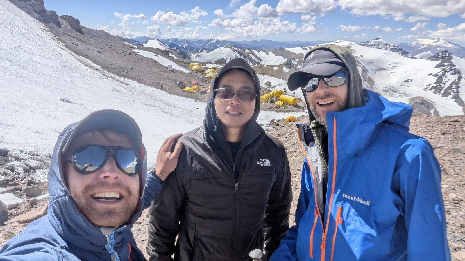 Three climbers at a high camp on Aconcagua with yellow tents, glacier and snow-covered peaks behind
