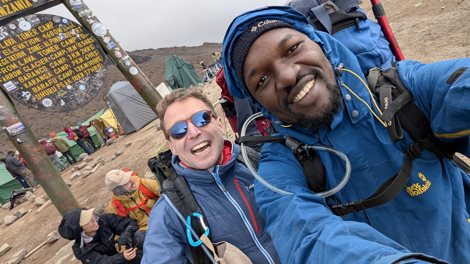 Sam and one of the Pomoja team grinning beside the Lava Tower Camp sign at 4,600 m, tents and the rest of the group behind