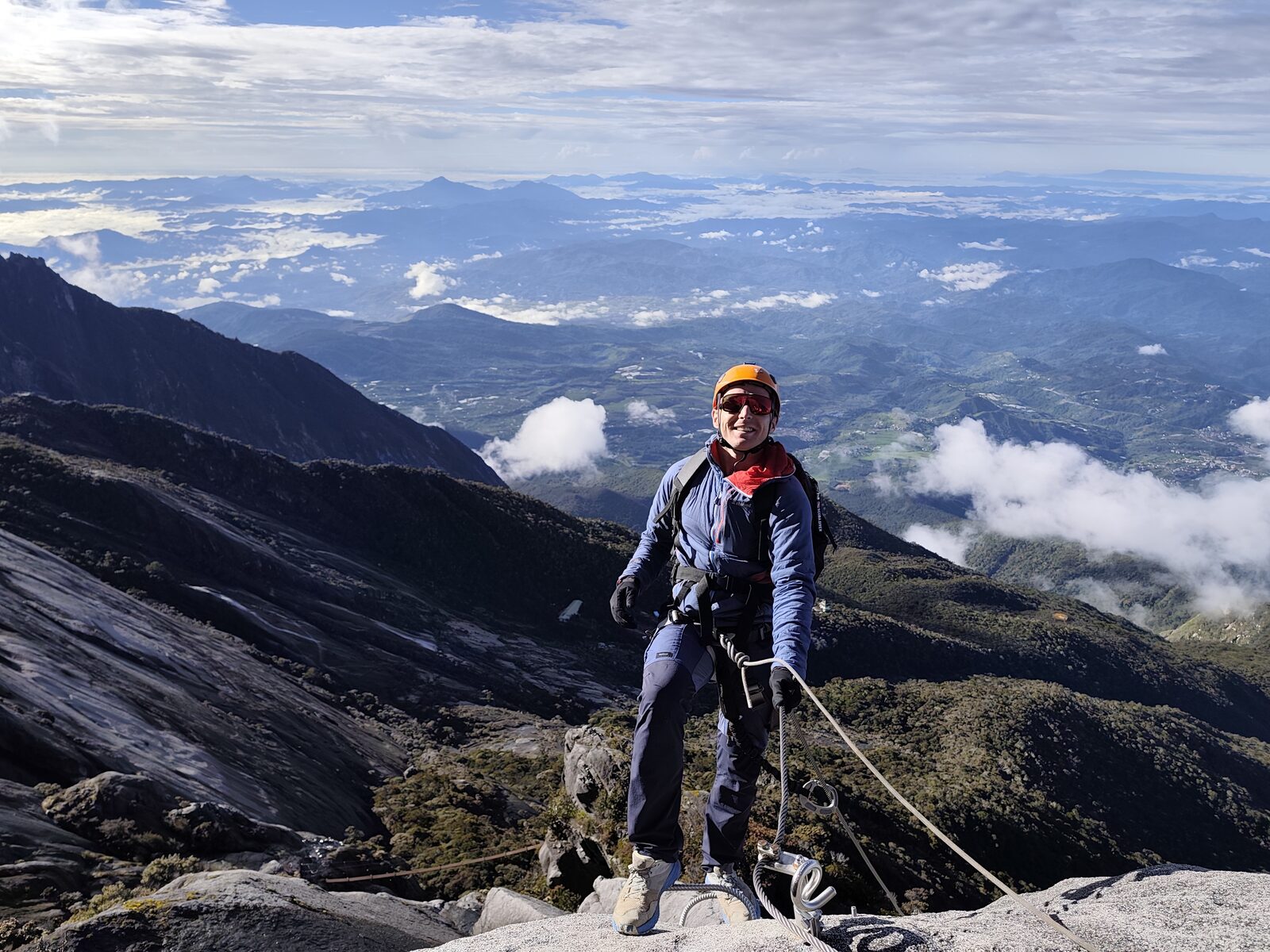 Sam on the via ferrata high on Mount Kinabalu, clipped to the cable, the whole of Sabah and a sea of cloud below